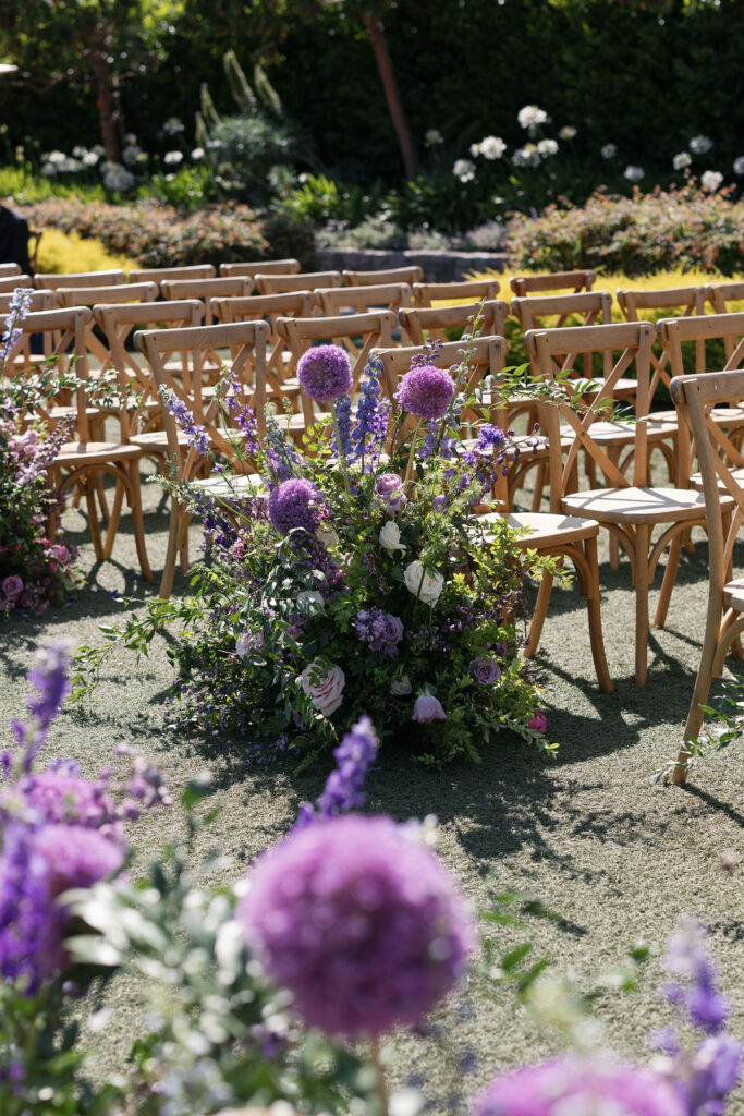 Rows of wooden chairs arranged outdoors with large purple and white floral arrangements along the aisle.