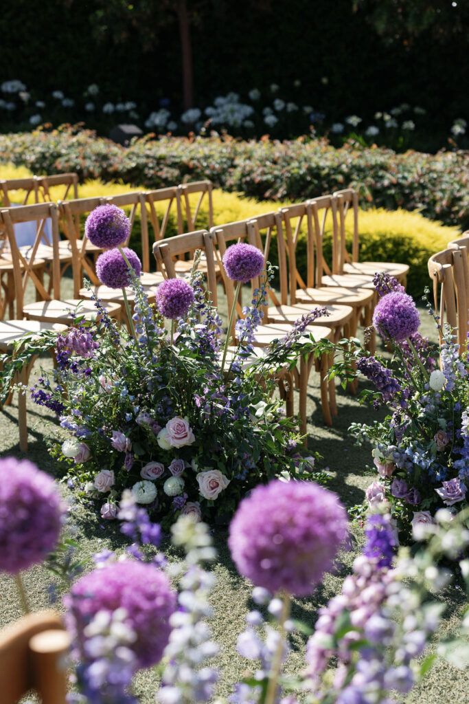Rows of wooden chairs are arranged outdoors beside large floral arrangements featuring purple and white flowers on a grassy lawn.