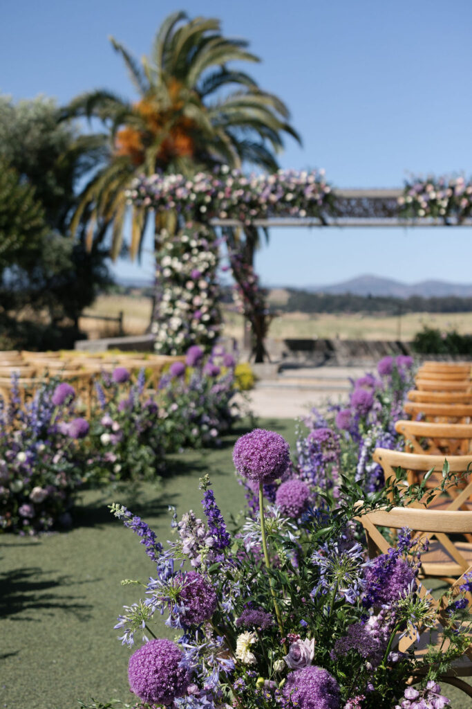 Outdoor wedding ceremony setup with wooden chairs and aisle lined with purple and lavender flowers; floral arch and palm tree in the background under a clear sky.