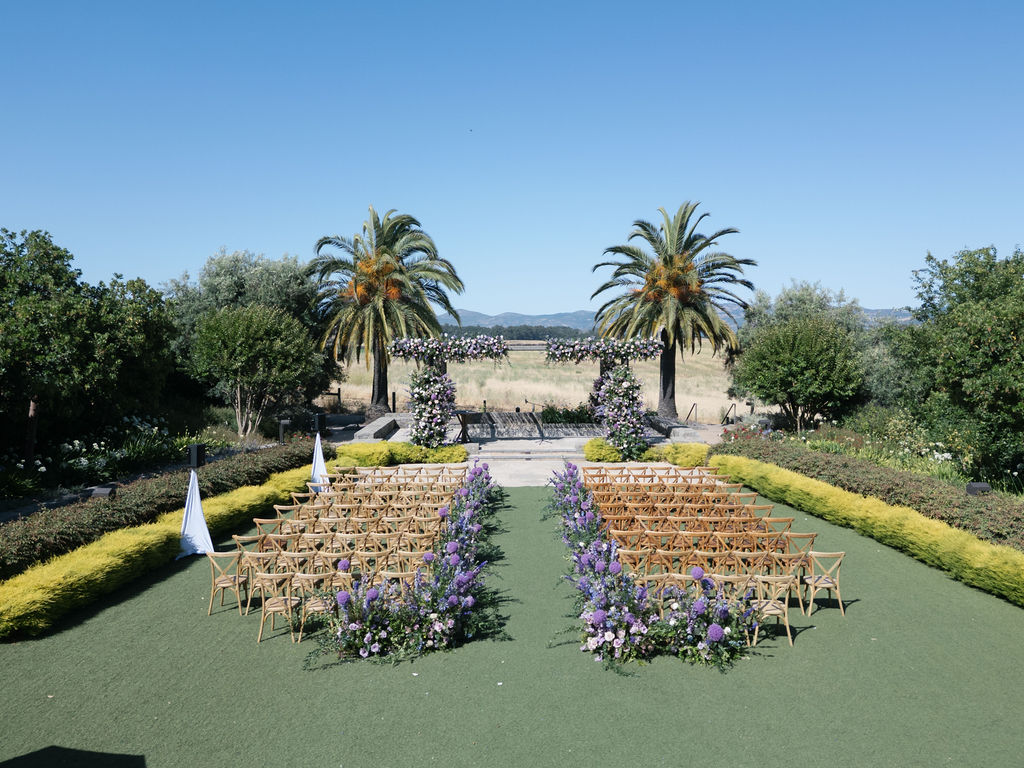 Outdoor wedding ceremony setup with rows of wooden chairs, purple floral arrangements along the aisle, two palm trees, and a floral arch under a clear blue sky.