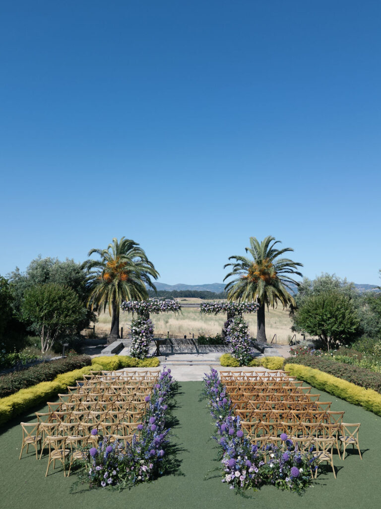 Outdoor wedding ceremony setup with rows of wooden chairs, purple floral arrangements, two palm trees, and a clear blue sky in the background.