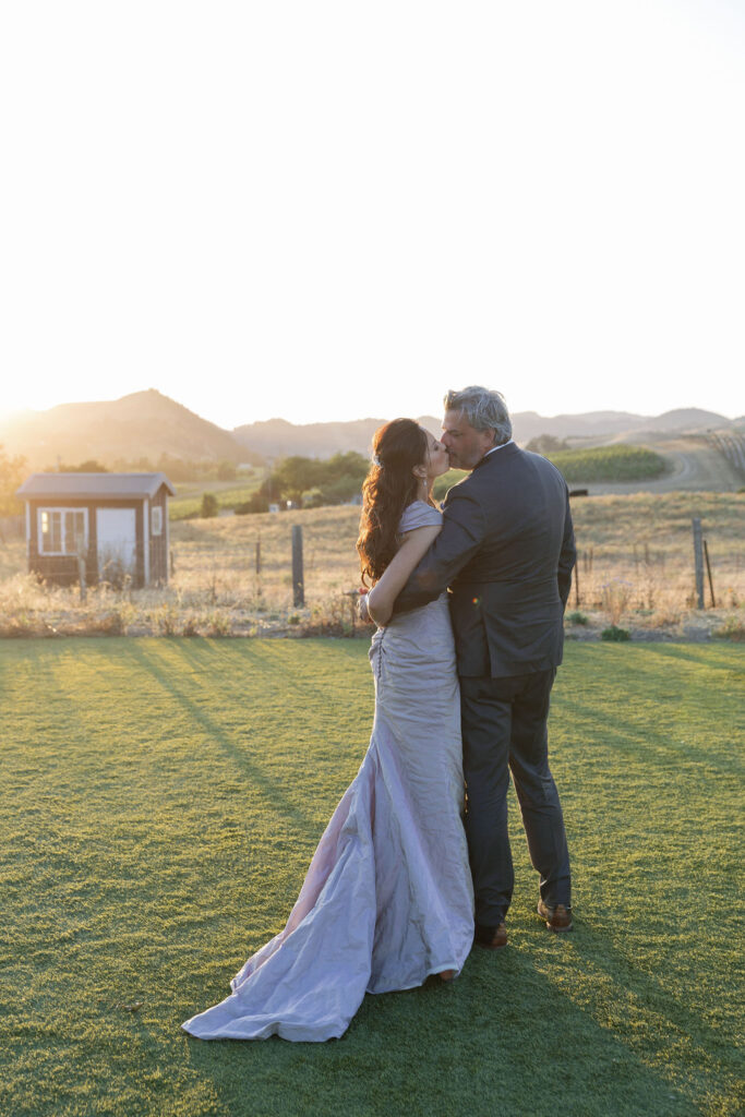 A couple dressed in formal attire stands close together on grass at sunset, facing away from the camera, with hills and a small building in the background.