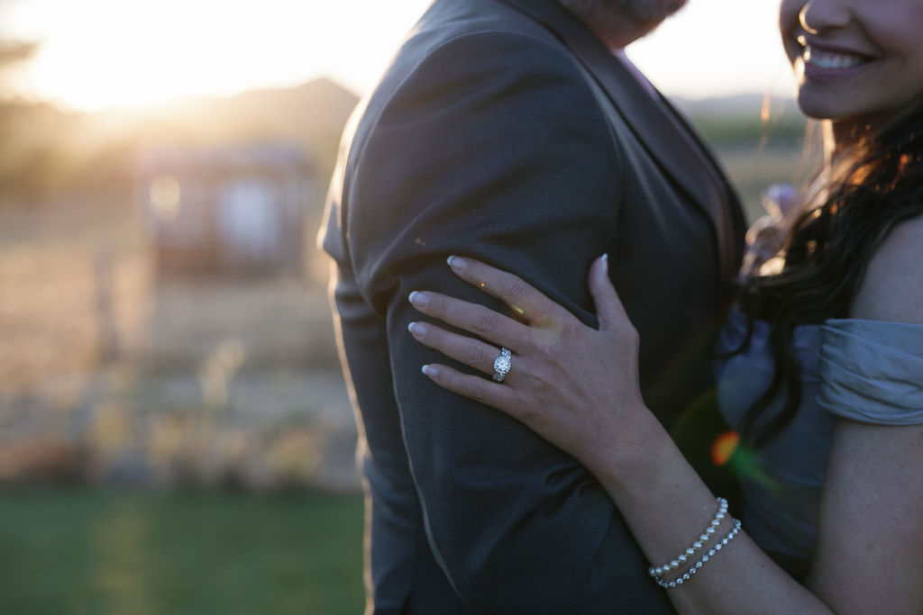 A woman with a pearl bracelet and an engagement ring embraces a man in a suit outdoors at sunset.