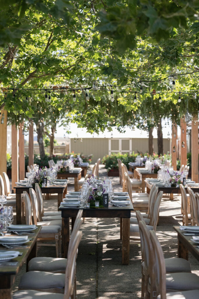 Outdoor dining area set for an event with wooden tables and chairs, floral centerpieces, and string lights under leafy trees.