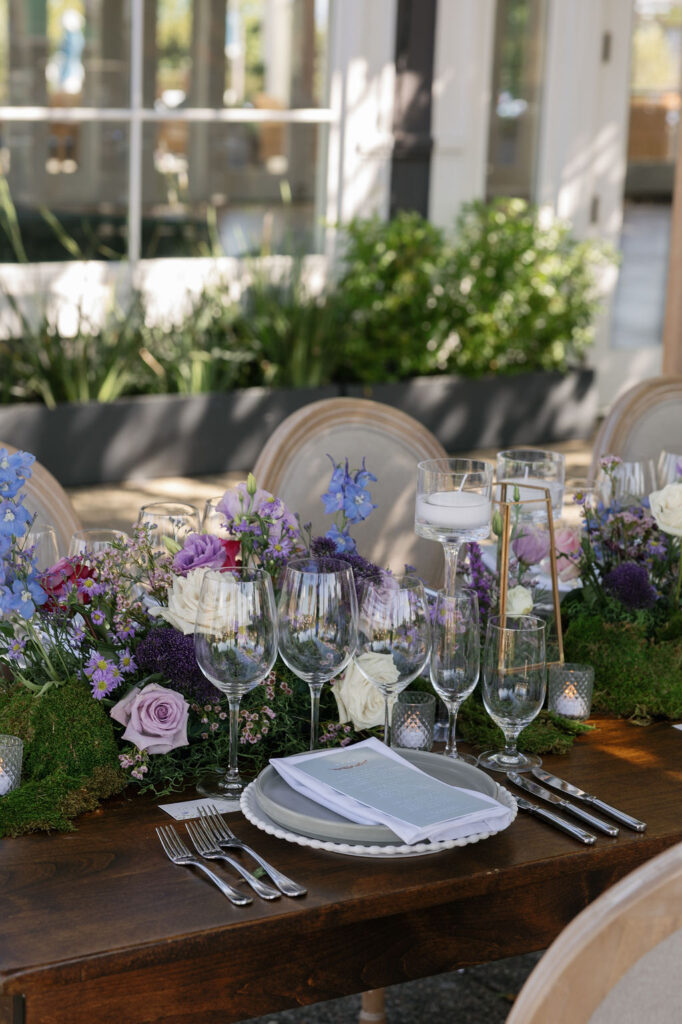 A decorated dining table set with glassware, plates, cutlery, and a floral centerpiece featuring purple, blue, and white flowers, in an outdoor setting.