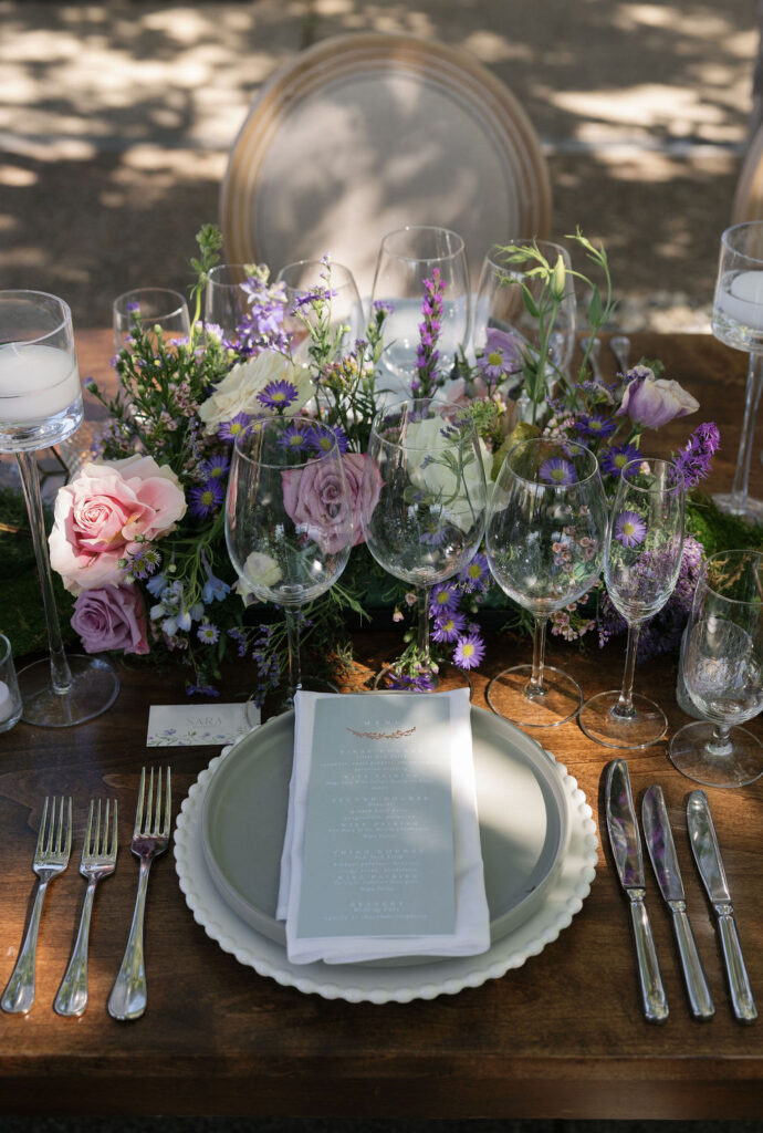 Elegant outdoor table setting with layered plates, silver cutlery, wine glasses, a printed menu, and a centerpiece of pink and purple flowers with greenery.