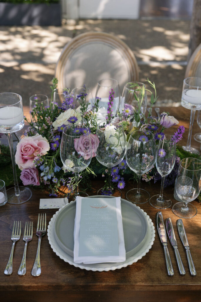 A formal table setting with multiple glasses, cutlery, a menu card, and a floral centerpiece with pink and purple flowers on a wooden table.