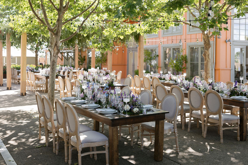 Outdoor dining area with multiple wooden tables and beige chairs set with floral centerpieces, plates, and glassware under string lights and leafy trees.