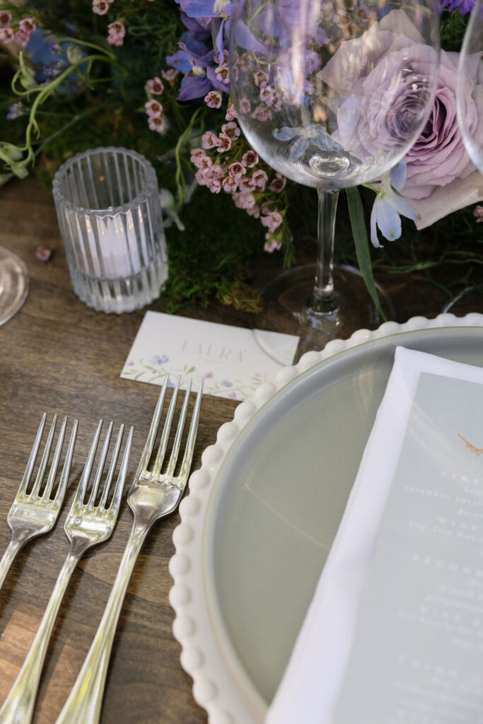 Close-up of an elegant table setting with four forks, a scalloped plate, a wine glass, a menu, a votive candle, and purple floral arrangement.