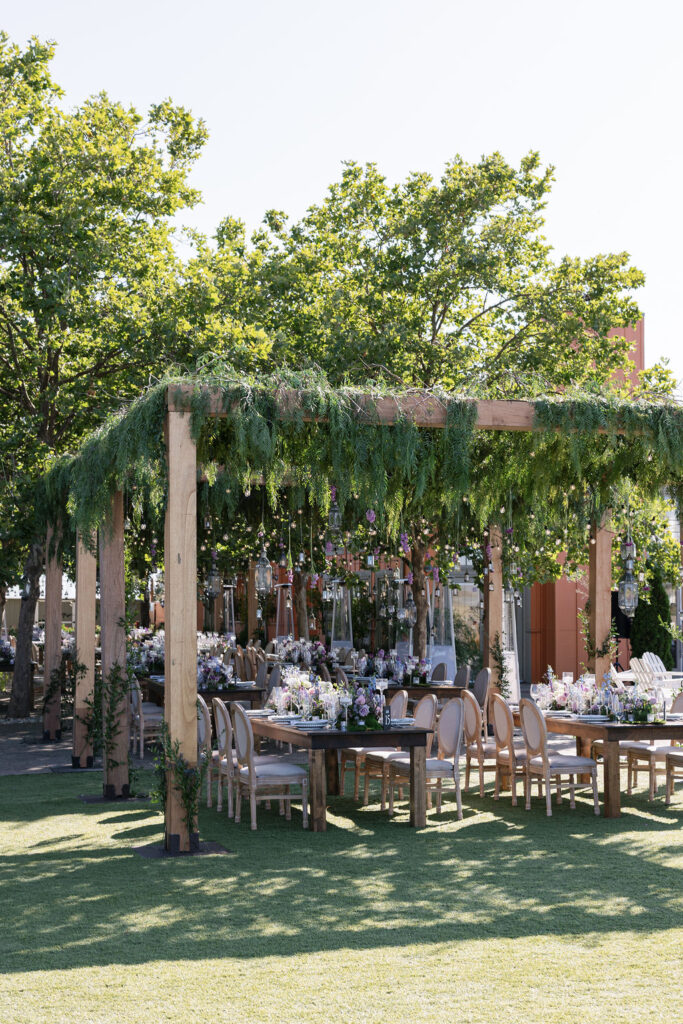 Outdoor dining area with wooden tables and chairs under a pergola covered in greenery, set up on a lawn with trees in the background.