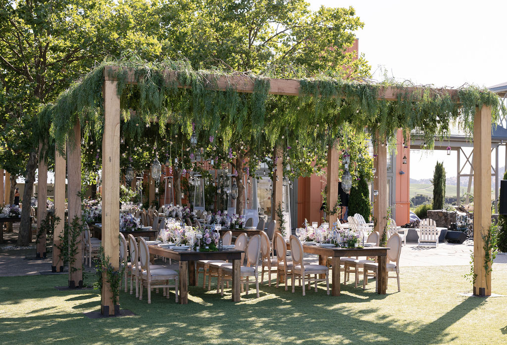 Long wooden tables and chairs are set up outdoors under a pergola covered with hanging greenery and surrounded by trees, prepared for an event or gathering.