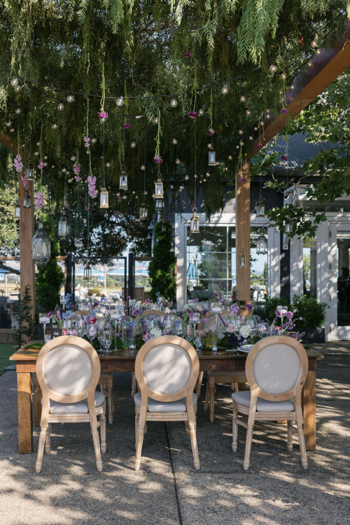 A wooden outdoor table set for a meal with six light-colored chairs, floral arrangements, and hanging glass lanterns under greenery.