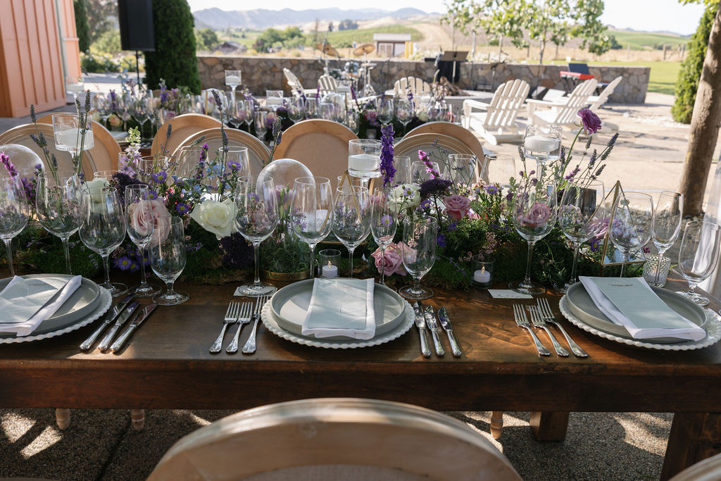 A decorated outdoor dining table set with plates, glasses, cutlery, menus, and floral centerpieces, with a scenic landscape in the background.