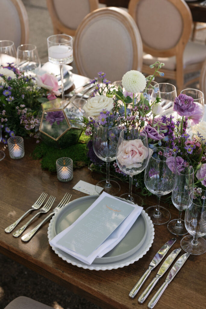 An elegant table setting with layered plates, multiple glasses, silver cutlery, a printed menu, candles, and a floral centerpiece featuring purple and white flowers.