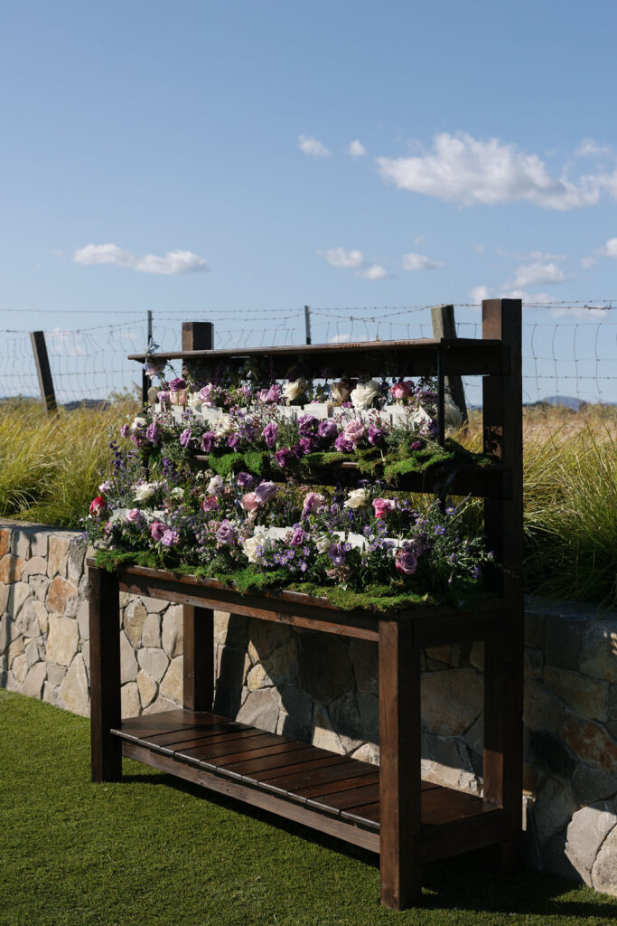 A wooden stand displays rows of purple and white flowers in front of a stone wall and wire fence under a blue sky with scattered clouds.