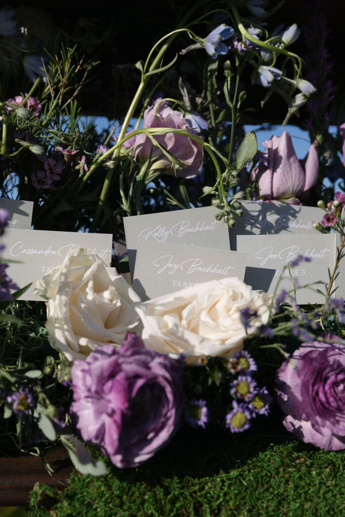 Close-up of name cards for table seating displayed among white and purple flowers and greenery.