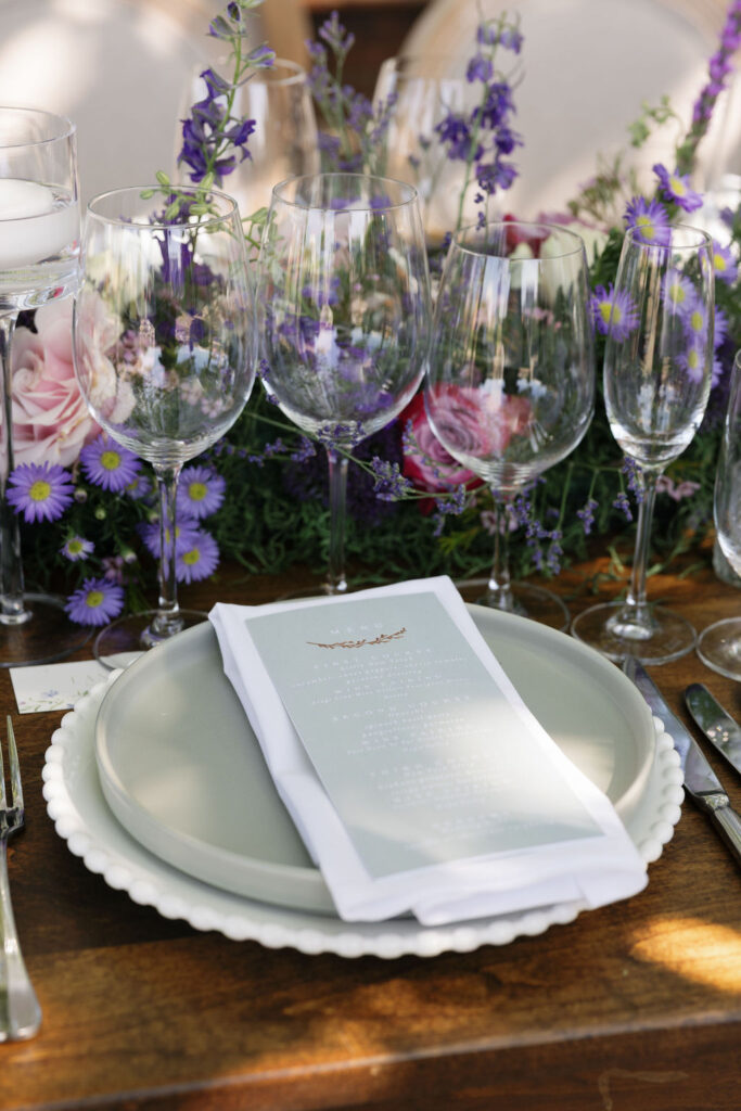 A formal table setting with a menu on a plate, surrounded by wine glasses, cutlery, and a floral centerpiece with purple and pink flowers.