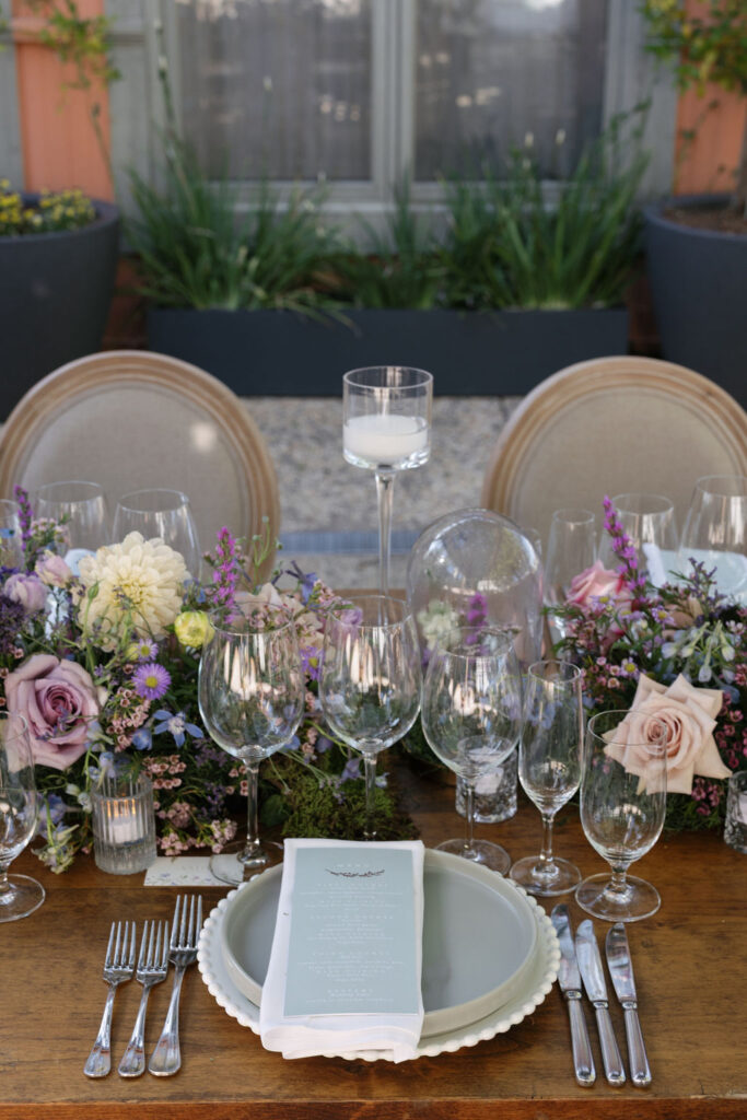 A formal outdoor table setting with multiple wine glasses, plates, silverware, a menu, and floral centerpieces featuring purple and pink flowers.