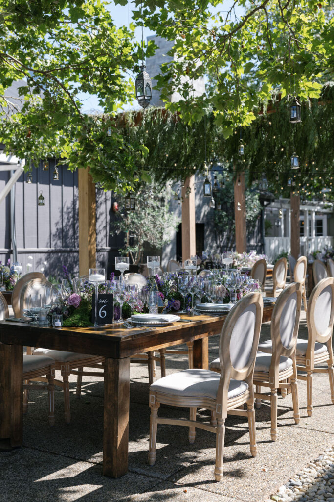 A long wooden outdoor dining table is set with glassware, floral centerpieces, and numbered cards, surrounded by cushioned chairs under hanging greenery and lanterns.