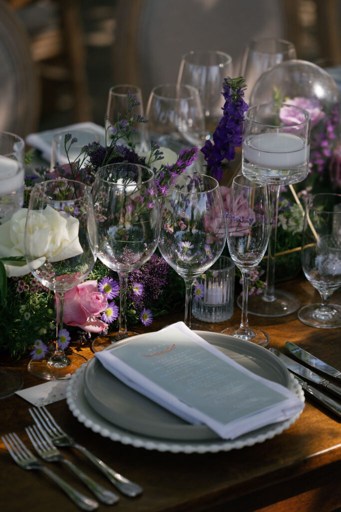 A formal table setting with plates, cutlery, several empty wine glasses, a printed menu, and a floral centerpiece with candles and purple, white, and pink flowers.