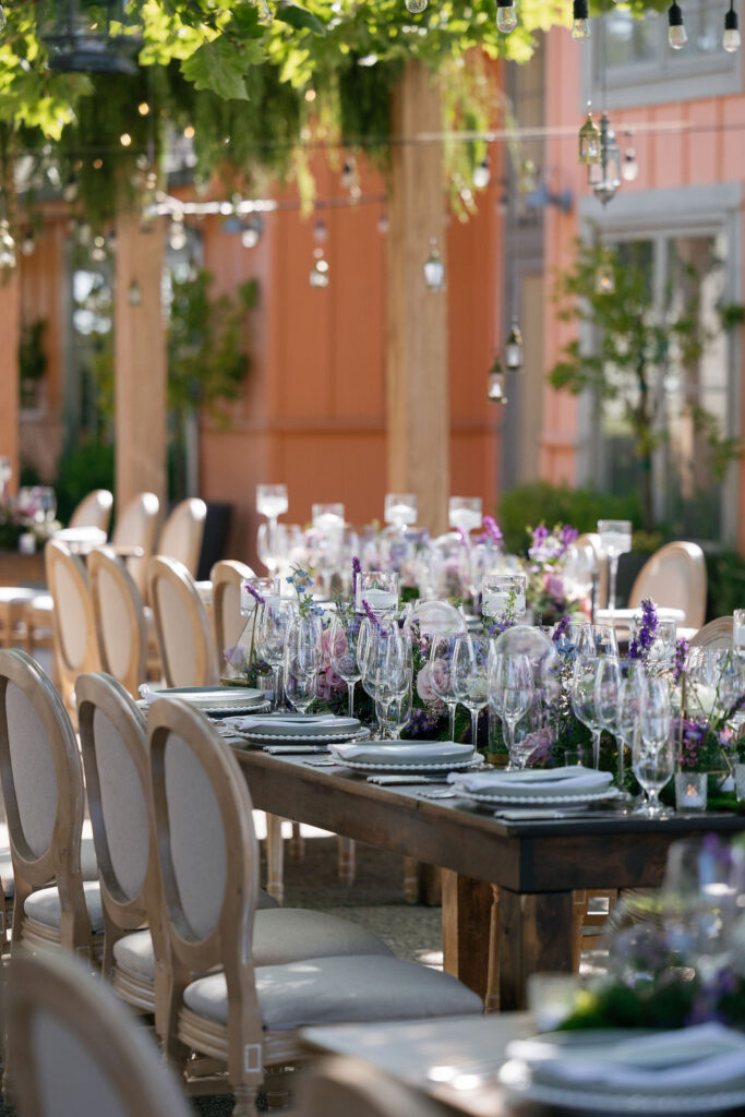 Elegant outdoor dining table set with glassware, plates, and floral centerpieces, surrounded by light-colored chairs under hanging string lights and greenery.