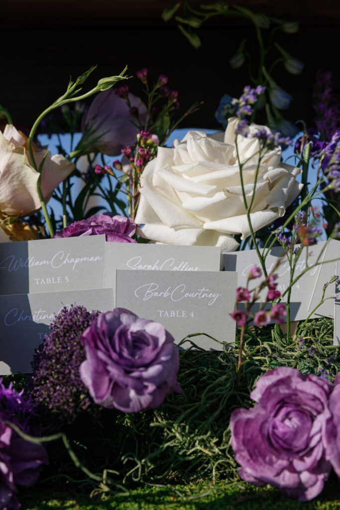 A close-up of floral arrangements with white and purple flowers, surrounded by several name cards placed in green moss.