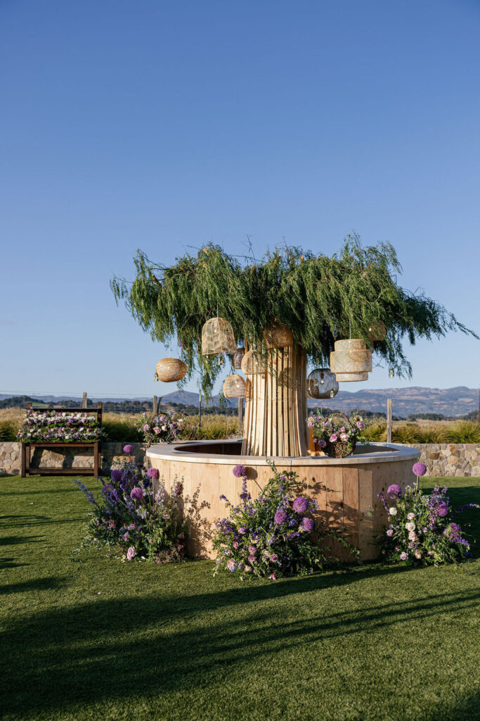 A large wooden circular structure with leafy branches and hanging woven lanterns, surrounded by purple and white flower arrangements on a grassy lawn.