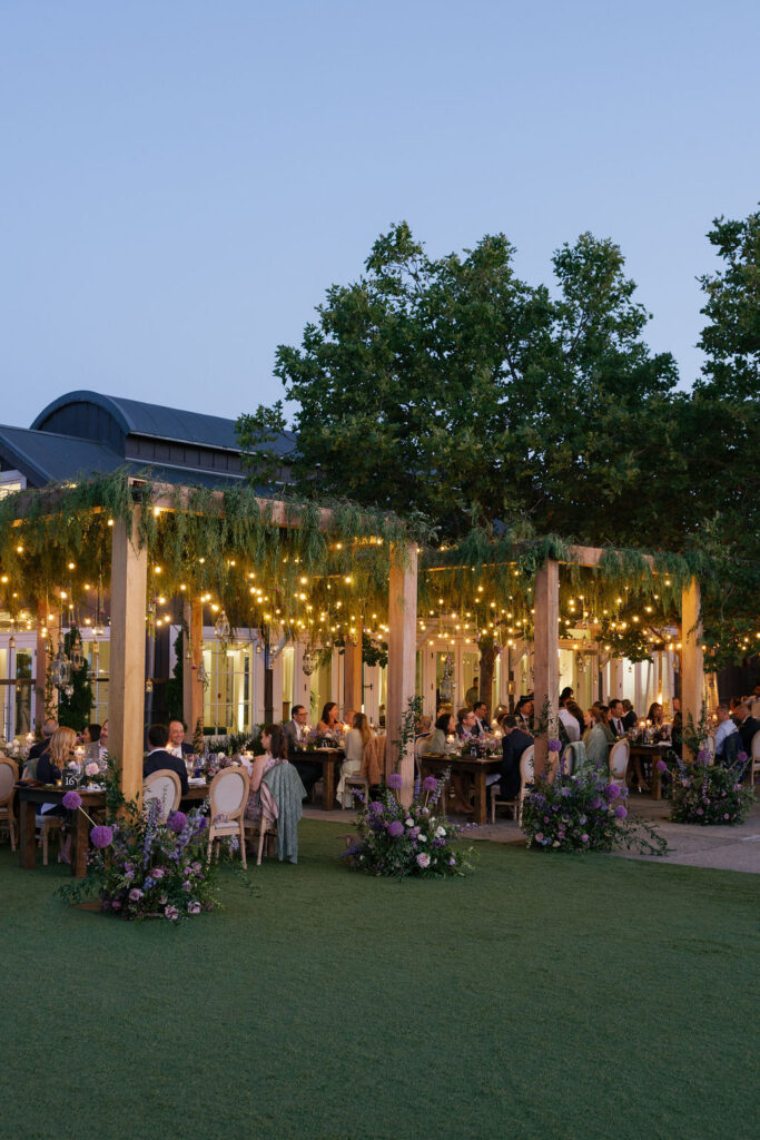 An outdoor evening event with people seated at tables under a pergola decorated with string lights, greenery, and flowers. Trees and a building are visible in the background.