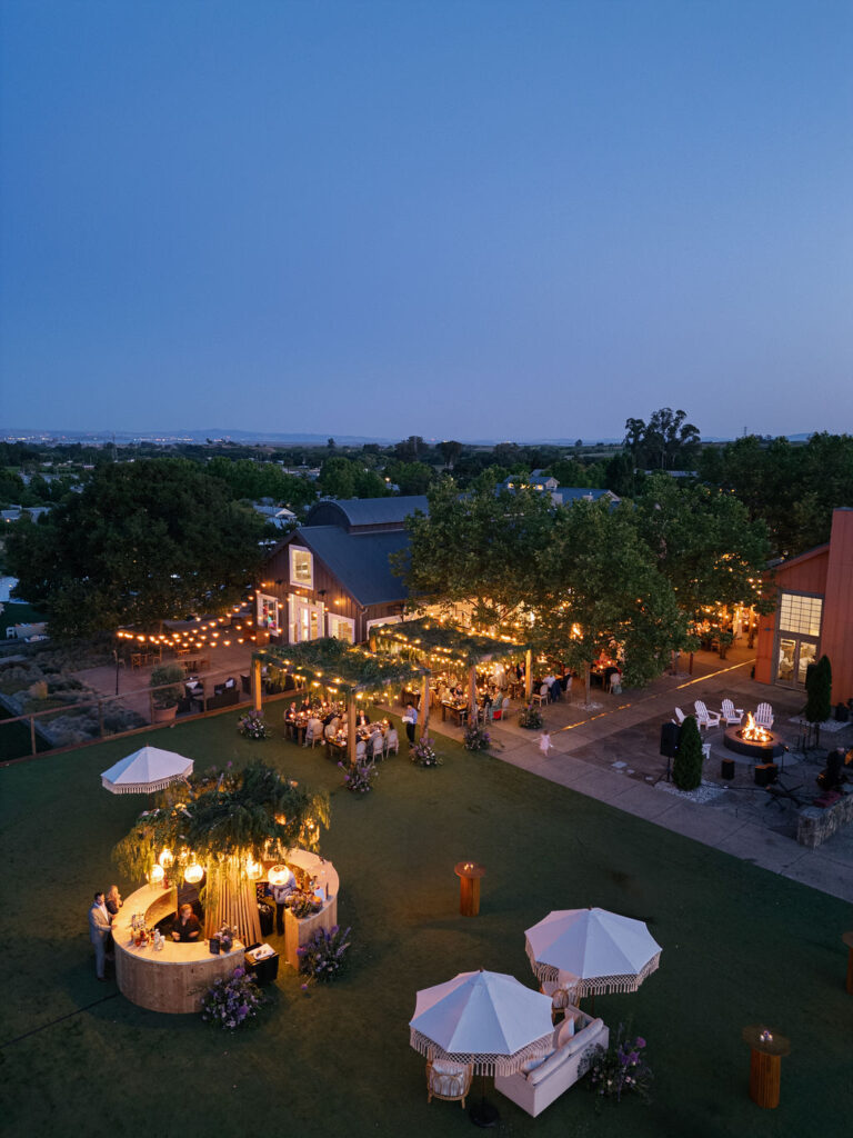 Outdoor evening event with string lights, guests mingling, decorated tables, lounge areas with umbrellas, and a rustic building surrounded by trees at dusk.