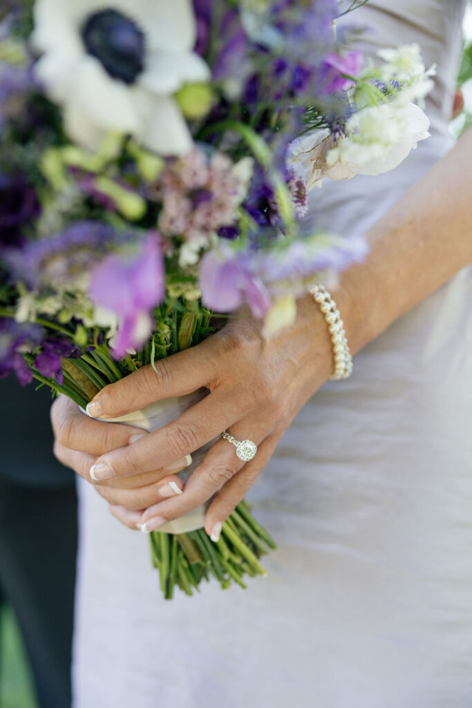 A person wearing a pearl bracelet and diamond ring holds a bouquet of purple and white flowers.