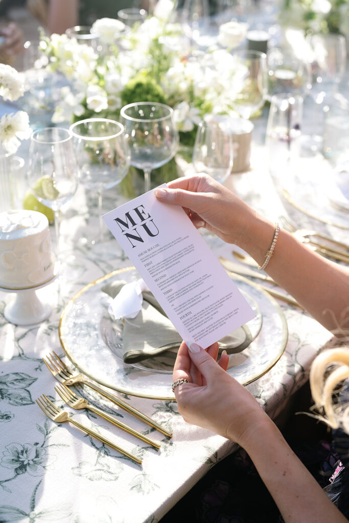 A person holding a printed menu at an elegantly set table with gold cutlery, glassware, and floral decorations.