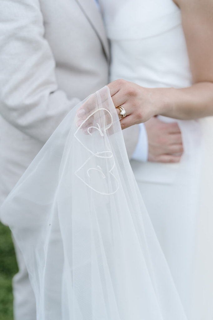 A bride holds a sheer veil with two embroidered hearts while standing close to a partner in a light suit.