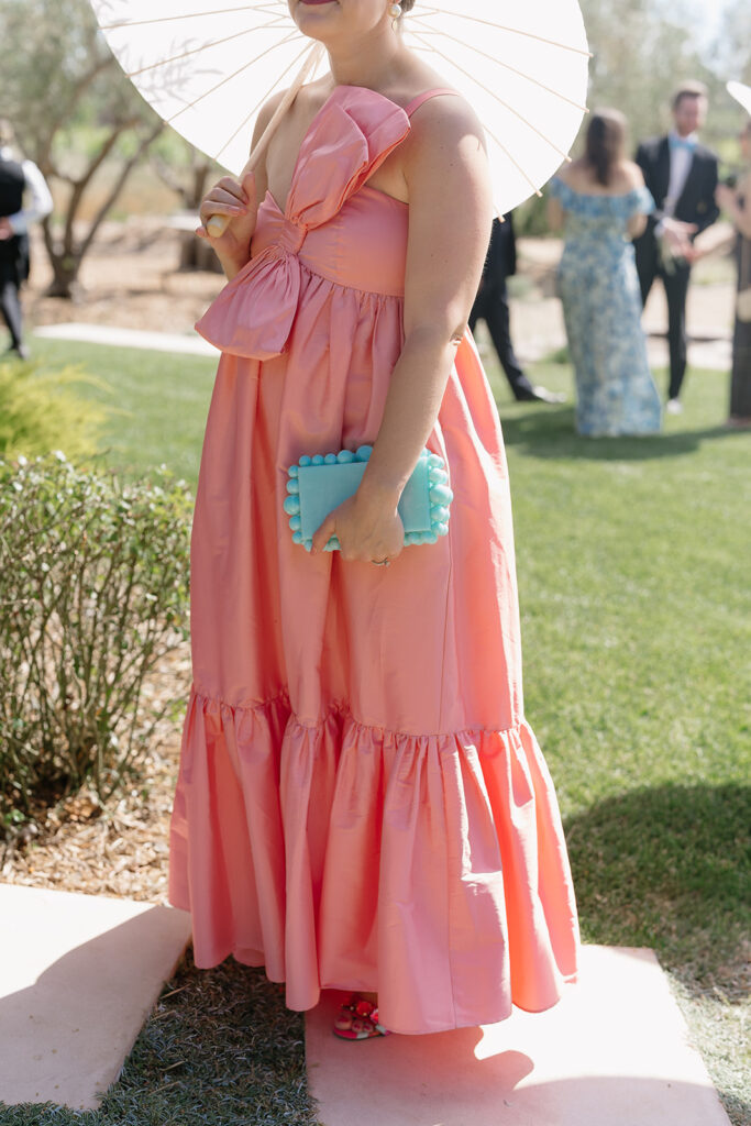 Woman in a pink dress holding a blue clutch and a white parasol stands outdoors on grass, with other people in formal attire in the background.