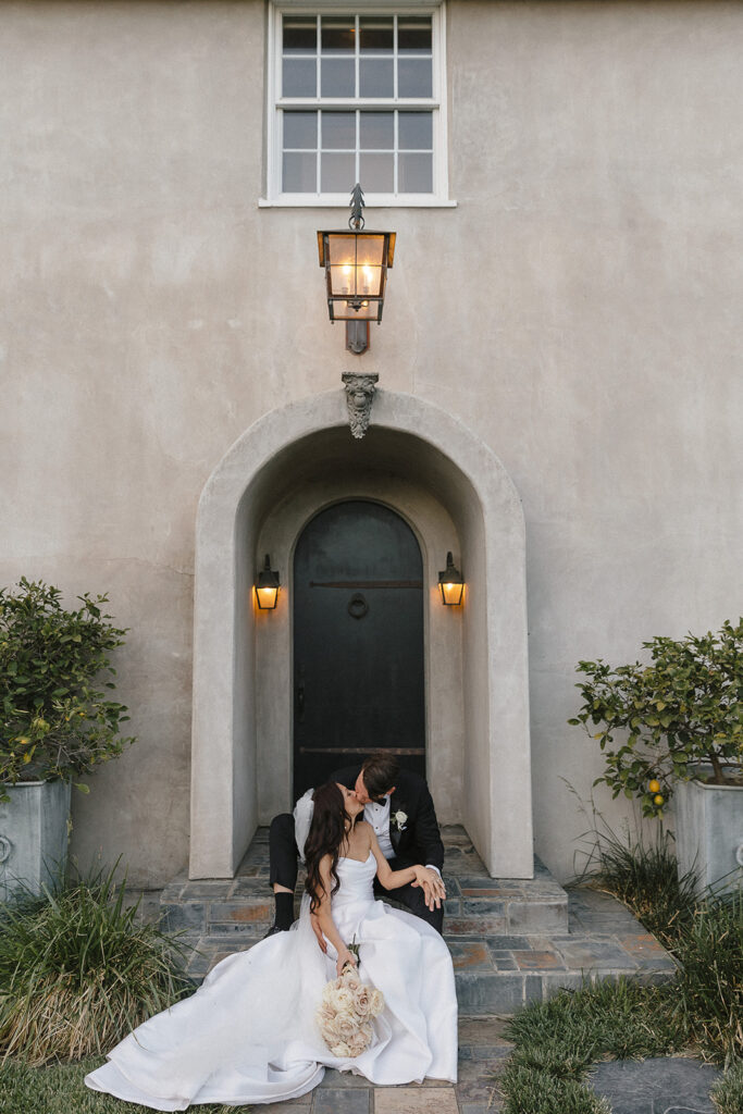 A bride and groom sit on steps outside a building, sharing an intimate moment. The bride holds a bouquet and both are dressed in wedding attire.