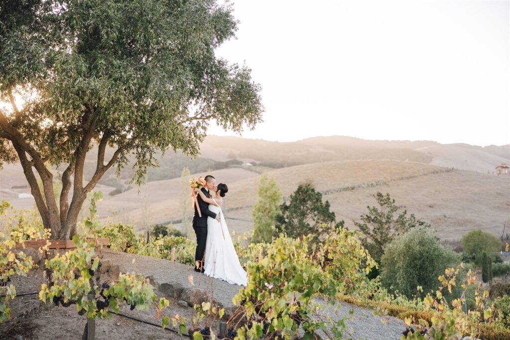 A bride and groom stand together on a vineyard path at sunset, surrounded by grapevines and hills in the background.