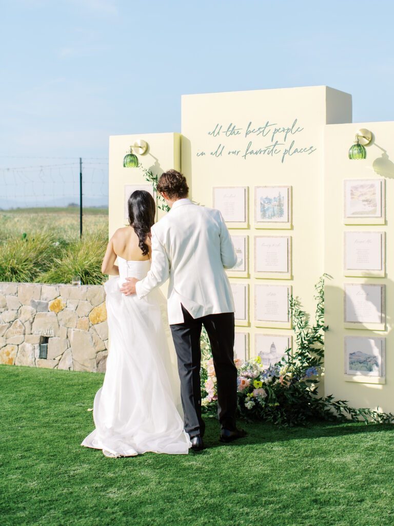 A bride and groom stand on grass in front of a cream-colored display board featuring framed papers and a sign that reads, "all the best people in all our favorite places.