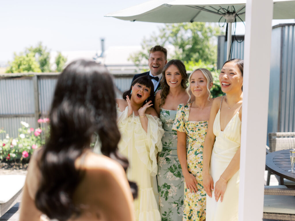 Five people, four women in pastel dresses and one man in a suit, pose and smile outdoors for a photo taken by a woman with long dark hair.