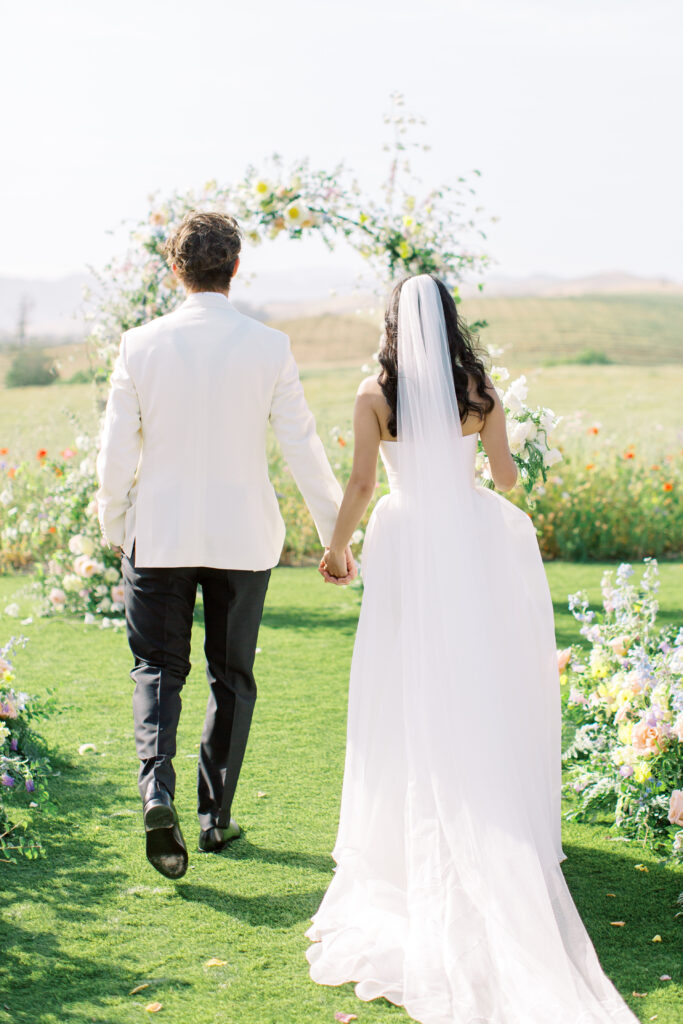 A bride and groom, seen from behind, walk hand in hand toward a floral wedding arch on a sunny outdoor lawn.