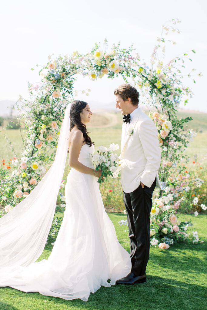 A bride and groom stand facing each other outdoors in front of a floral arch, dressed in wedding attire, on a sunny day.