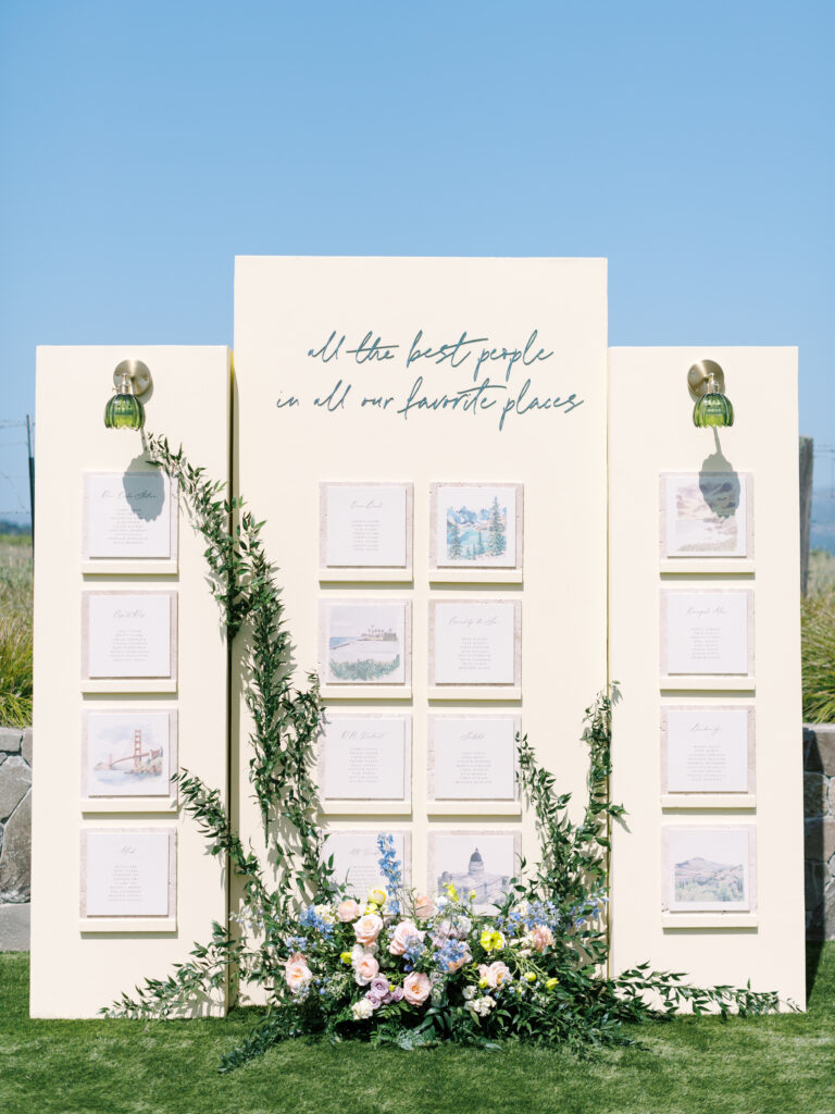 A cream-colored seating chart display with handwritten calligraphy, framed cards featuring illustrations, greenery, and pastel flowers arranged at the base, set outdoors under a clear sky.