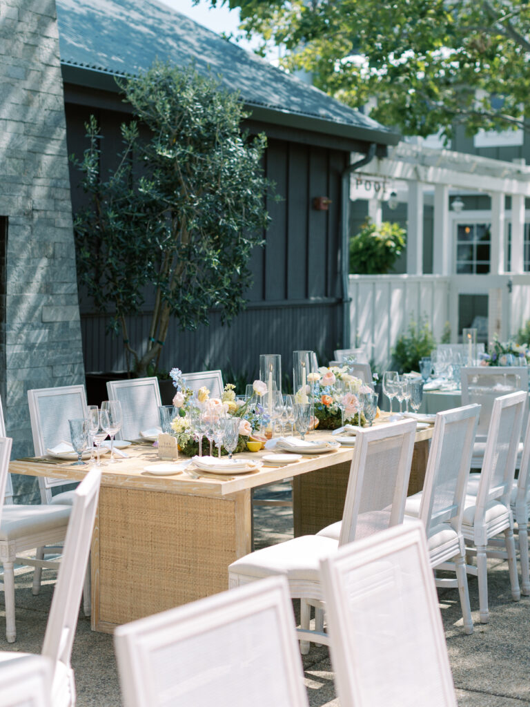 Outdoor dining table with white chairs, set with glassware, floral centerpieces, and candles, positioned next to a dark building and greenery on a sunny day.