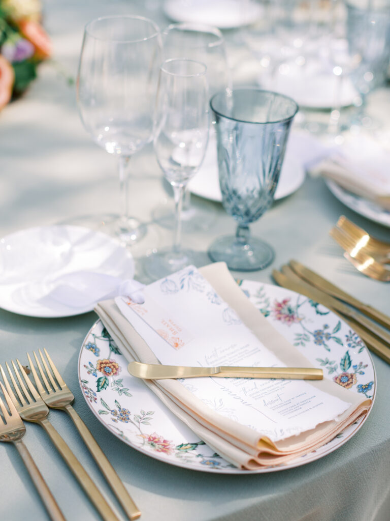 A floral-patterned plate with a beige napkin, a printed menu, and gold flatware are set on an elegant table with glassware.