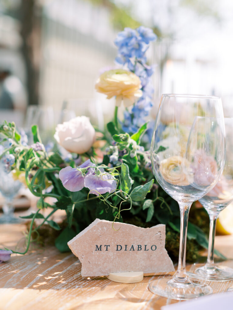 A stone place card labeled "MT DIABLO" sits on a table next to empty wine glasses and a floral arrangement with pastel flowers.