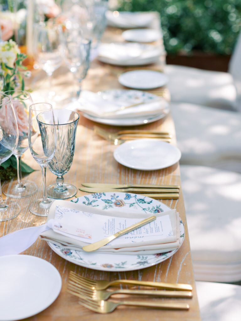A wooden table set with floral plates, gold cutlery, wine glasses, folded napkins, and printed menus, with cushioned chairs and floral centerpieces in the background.