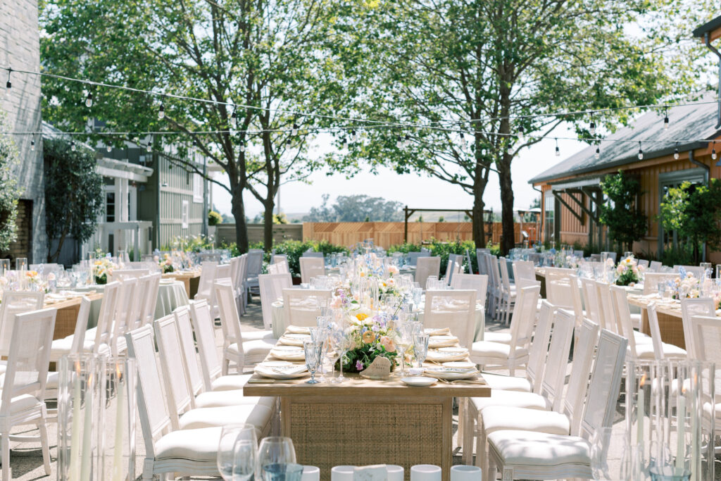 Outdoor event setup with long dining tables and white chairs arranged under string lights, featuring floral centerpieces and place settings, surrounded by buildings and leafy trees.