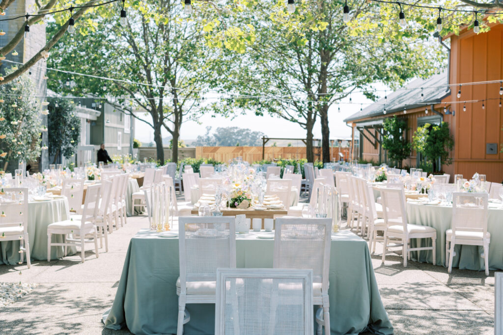 Outdoor event setup with white chairs and pastel tablecloths under string lights and trees, tables set with plates and centerpieces, in a courtyard with surrounding buildings.