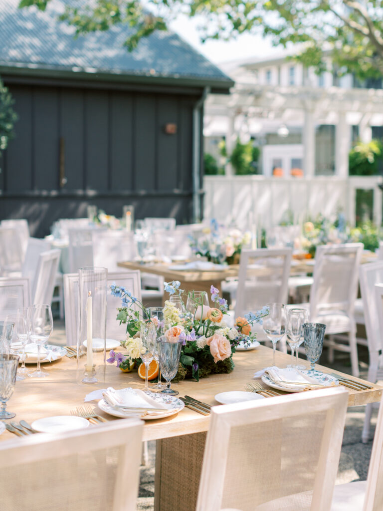 Outdoor dining tables set with floral centerpieces, glassware, and place settings, surrounded by white chairs in a garden setting.