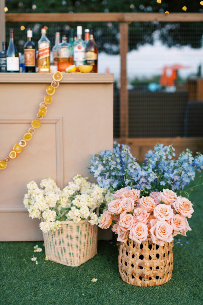 A bar counter decorated with dried orange slices, bottles of liquor on top, and two baskets of flowers—one with white blooms, the other with pink roses and blue flowers—on artificial grass.