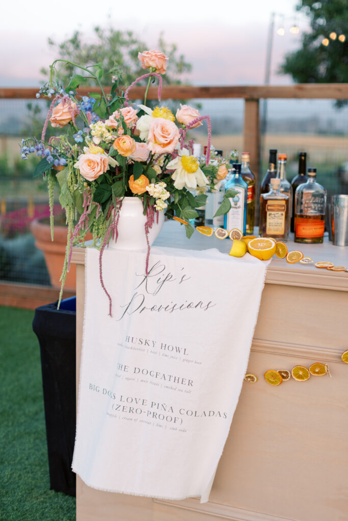 A bar setup outdoors features assorted liquor bottles, a floral arrangement, dried citrus decor, and a towel listing specialty drinks named after dogs.