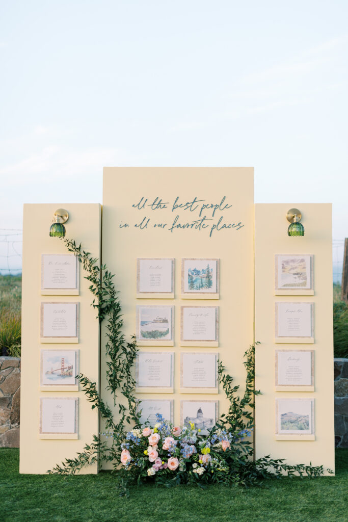 A yellow display board with framed illustrations and text, decorated with greenery and flowers, stands outdoors. Top text reads: “All the best people in all our favorite places.”.